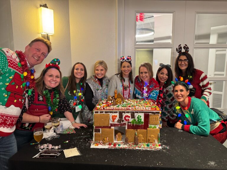A group of nine adults in festive sweaters pose around a decorated gingerbread house on a table, with holiday lights and decorations visible.