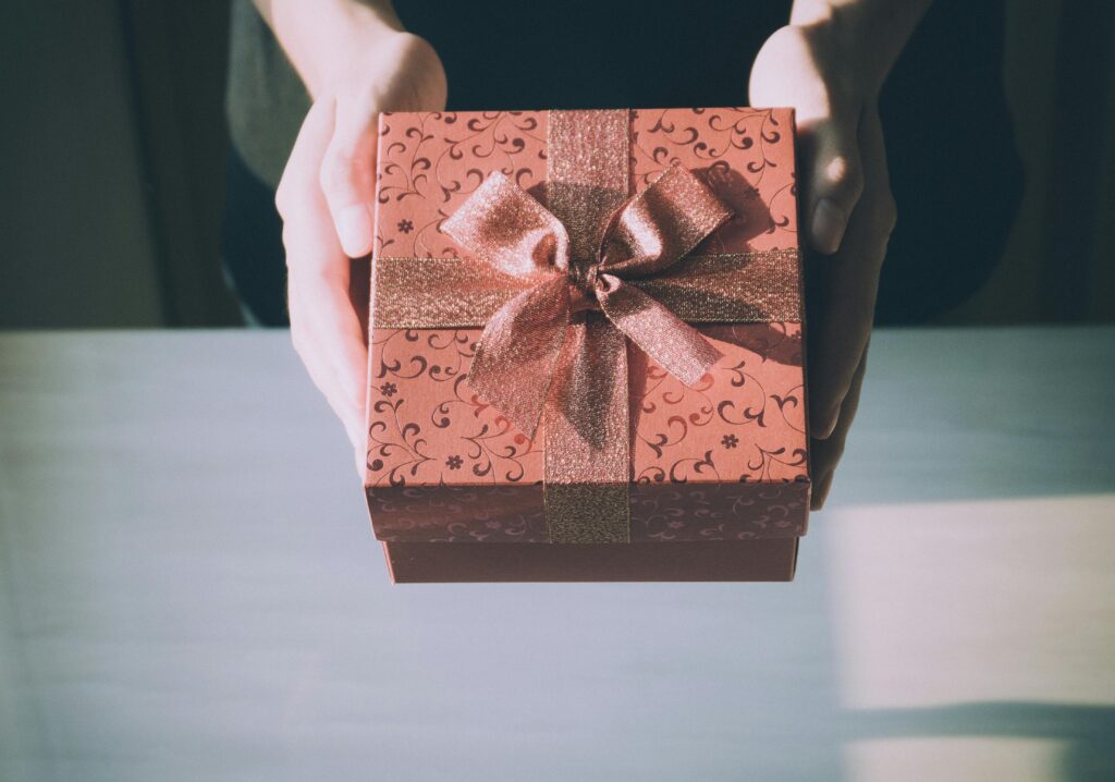 Hands holding a red gift box with a patterned design and a glittery ribbon bow, over a light-colored table.