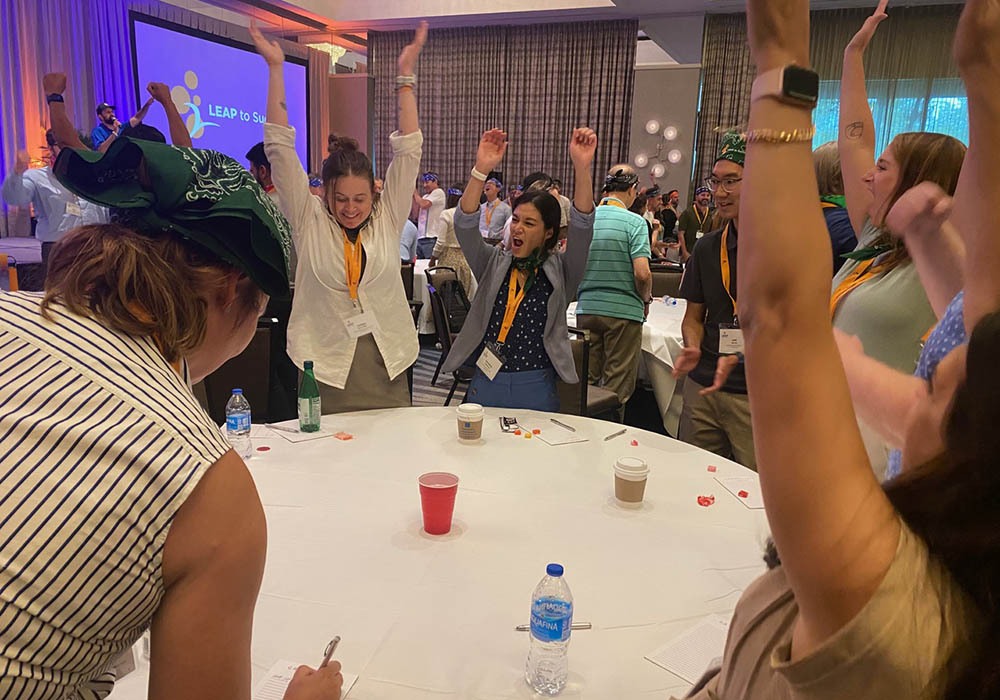 People enthusiastically celebrating around a table at an indoor event, with some raising their hands. A screen in the background reads "LEAP to Success." Various drinks are on the table.