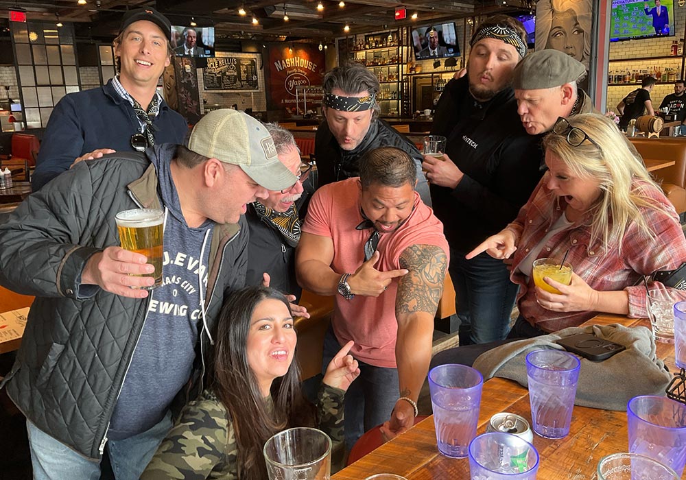 A group of people gathered in a bar with expressions of laughter and surprise, some holding drinks.