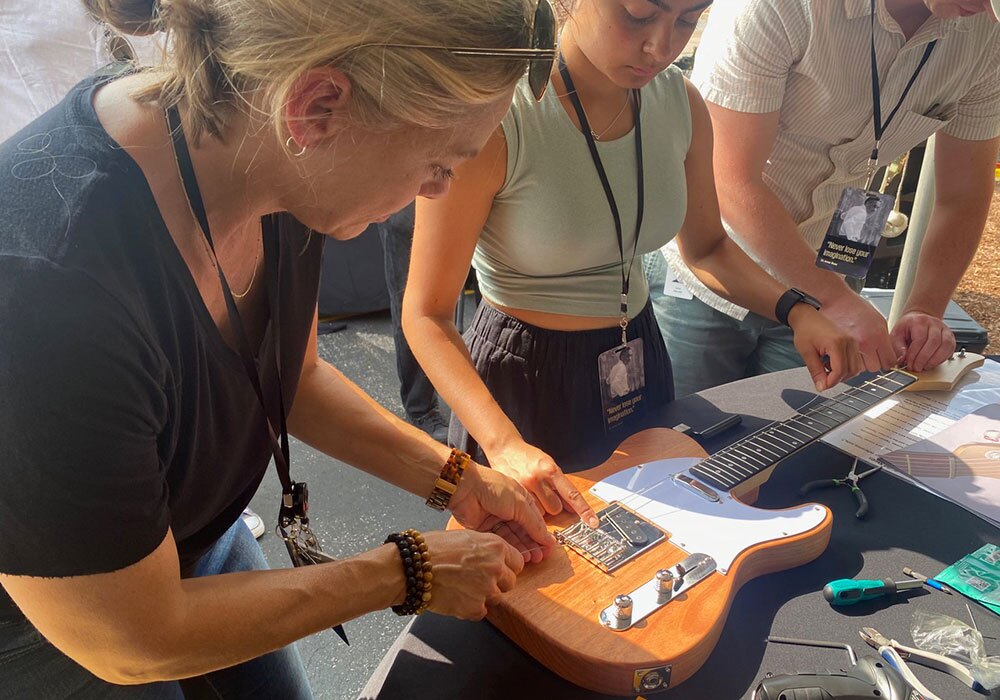 Three individuals are working together to set up an electric guitar on a table, with tools and guitar components visible around them.