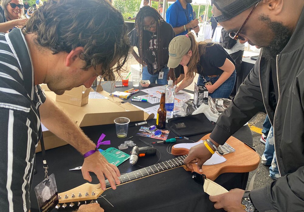 Two men assemble an electric guitar at a table, while a woman in the background sorts through papers. Various tools and drink containers are on the table.