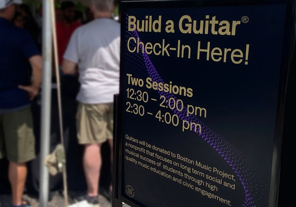 Sign reads: "Build a Guitar Check-In Here! Two Sessions: 12:30 - 2:00 PM, 2:30 - 4:00 PM." Event donates guitars to Boston Music Project. Blurred background of people standing.