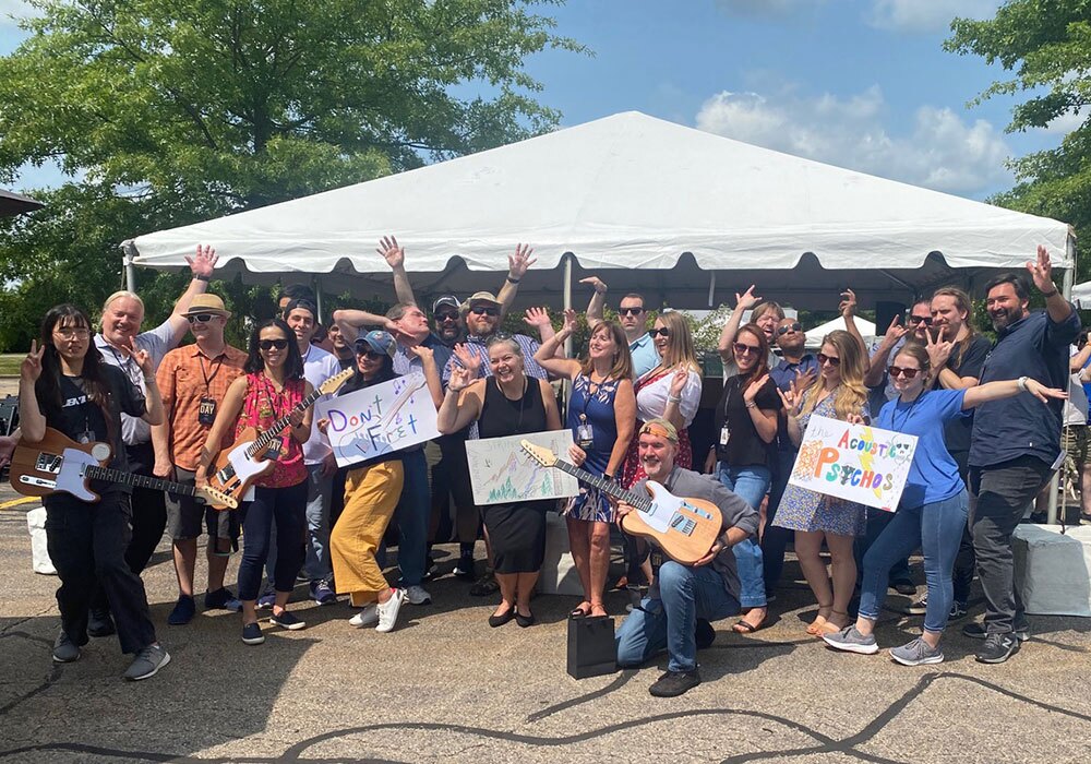 A group of people posing outdoors under a white tent, some holding guitars and signs, wearing casual clothing, with trees in the background.