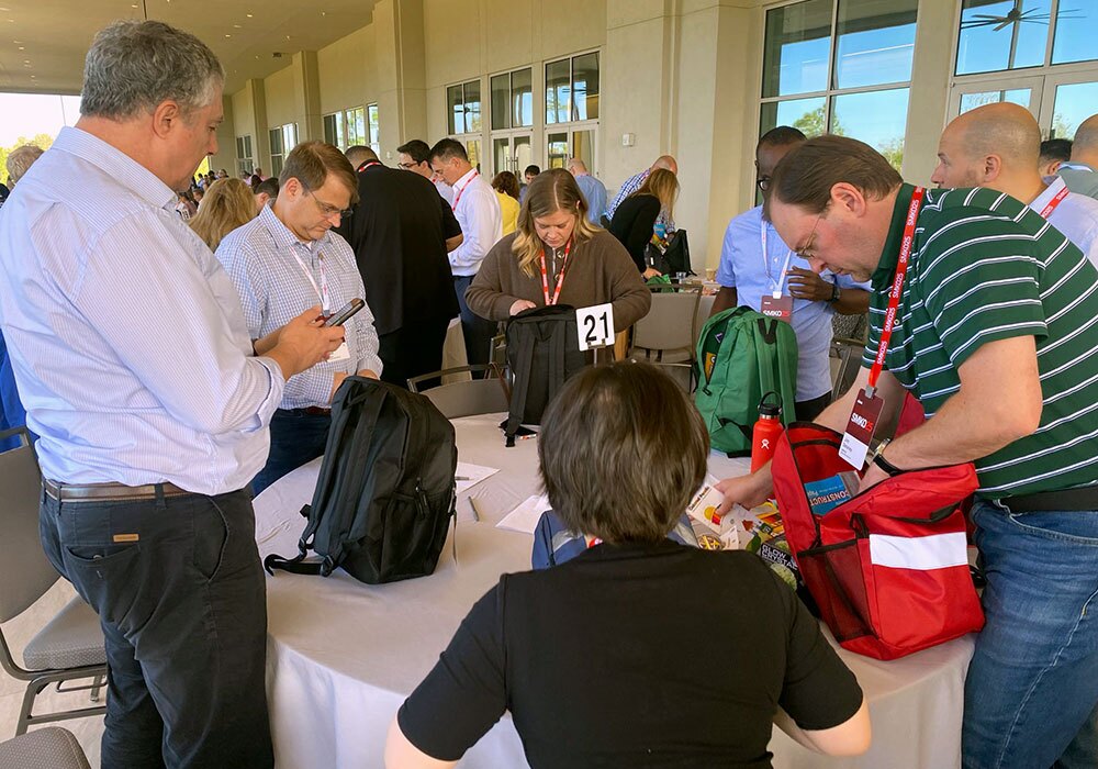 People standing around a table, looking into backpacks, at an indoor event with large windows.