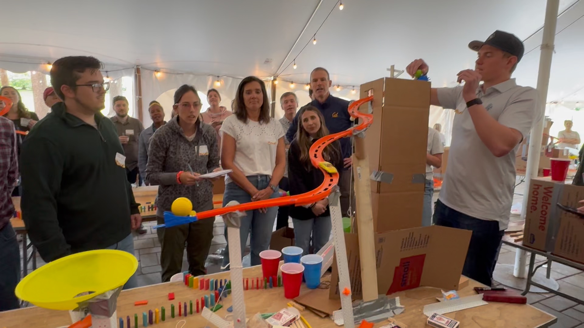 A group of people watch as a person adjusts a marble run made from cardboard, tracks, and dominoes at an indoor event.