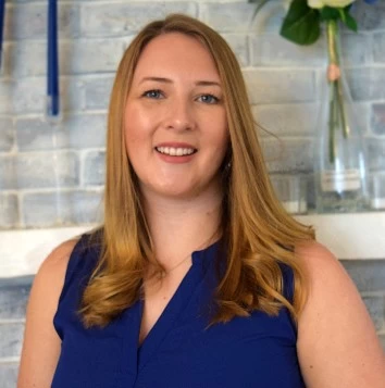 A woman in a blue top standing in front of a fireplace.