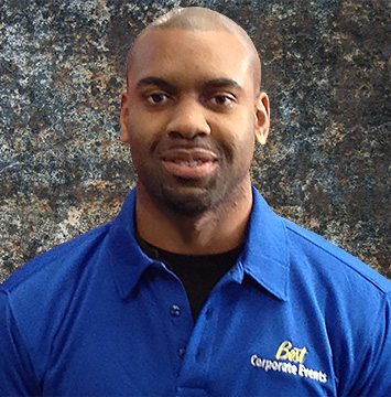 A black man in a blue shirt standing in front of a wall.