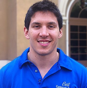 A young man in a blue shirt smiling in front of a building.