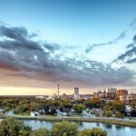 Panoramic view of a cityscape with a river and trees in the foreground, buildings and skyline in the background, and a sky with clouds during sunset.