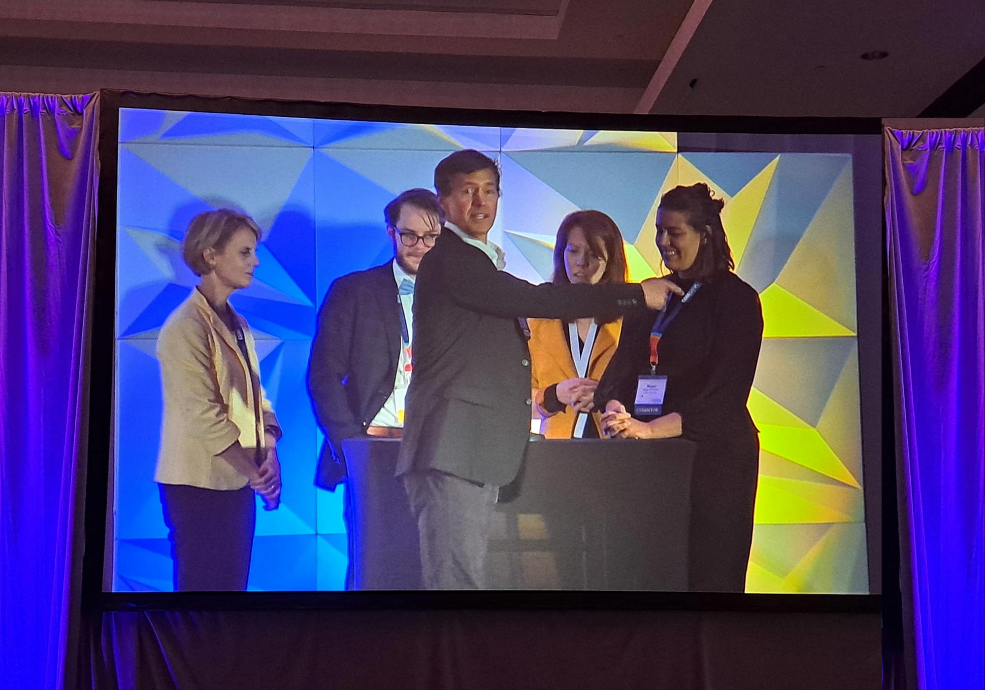 A group of five people stands on a stage with a geometric blue and yellow background. One person in a grey suit speaks while the others listen attentively. Bright lighting illuminates the scene.
