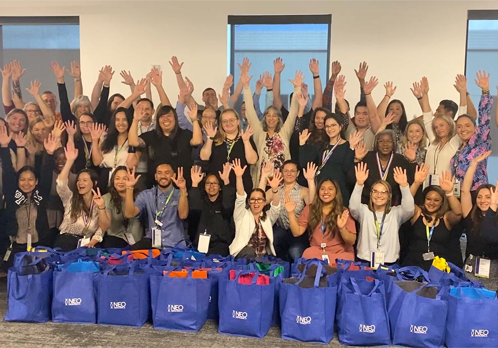 A large group of people poses with raised hands, sitting and standing behind a row of colorful cancer care packs in an office setting.