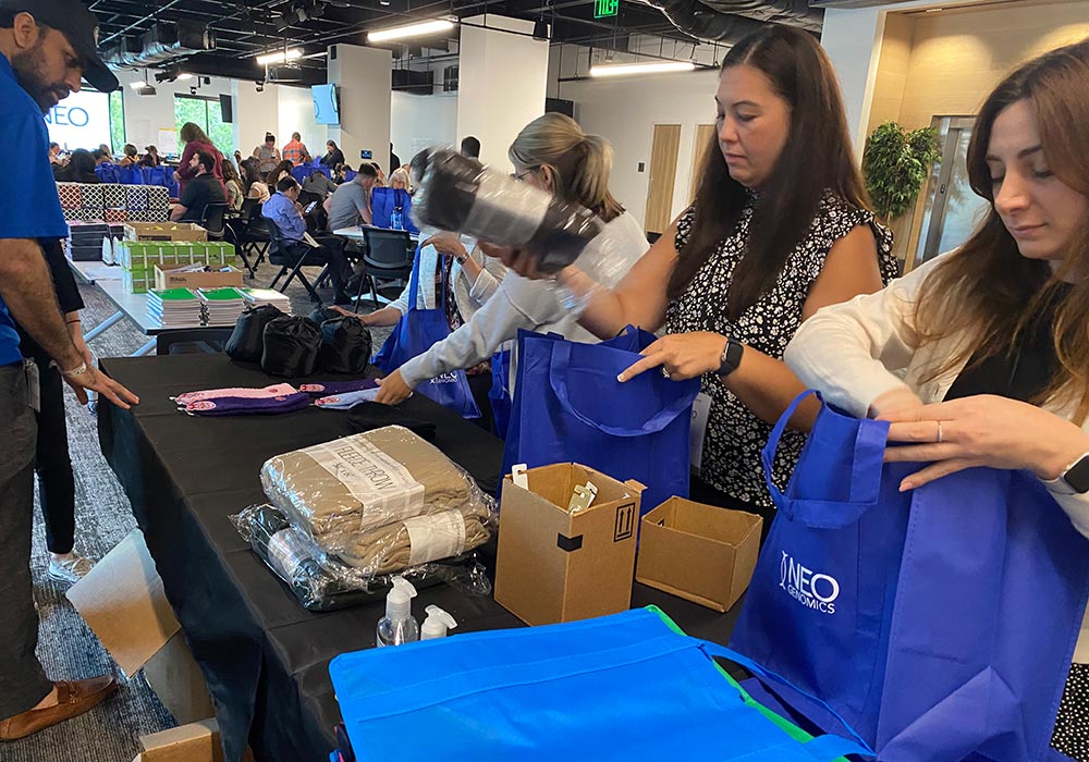 People are packing various items, including blankets and sanitizers, into blue cancer care packs at a table in an office setting. Other individuals are seen seated and working in the background.