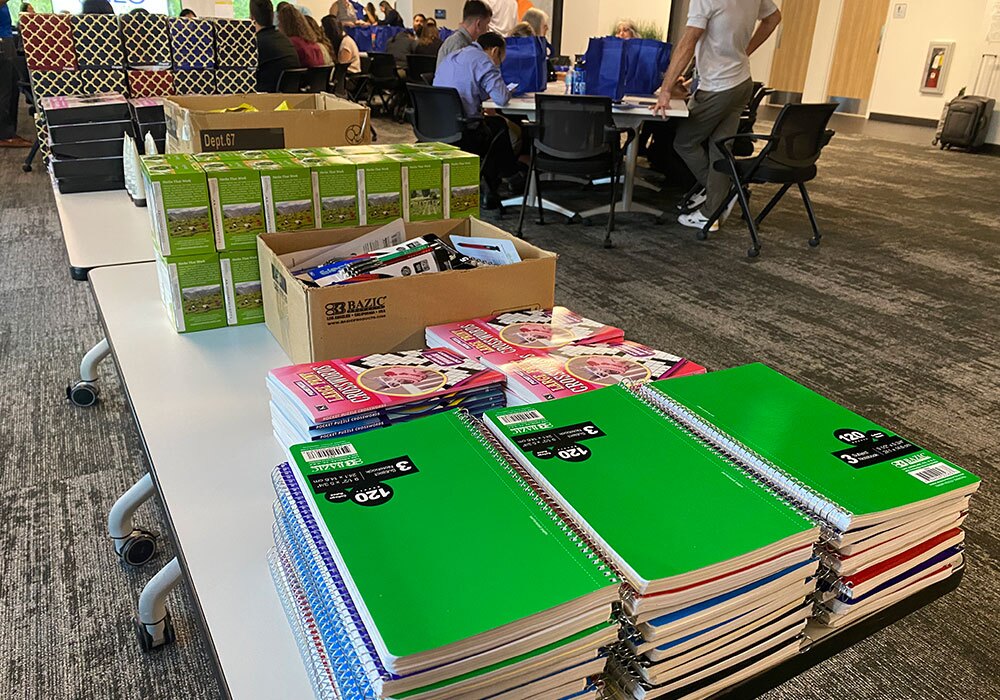 A table filled with stacks of spiral notebooks, packs of highlighters, pens, and other stationery items. People are sitting and organizing supplies in the background, including thoughtful cancer care packs for those in need.