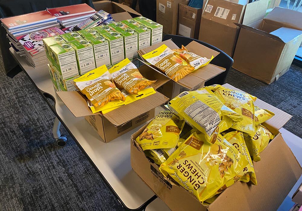 Boxes of fruit juice packs and ginger chews are displayed on a table, with cancer care packs and school notebooks in the background.