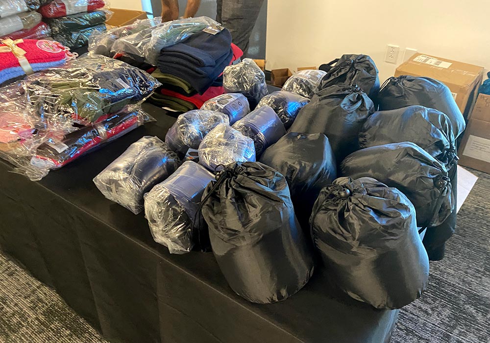 A table is filled with neatly arranged winter clothing items, including folded sweaters, rolled sleeping bags, and plastic-wrapped hats, all prepared as part of cancer care packs for distribution.