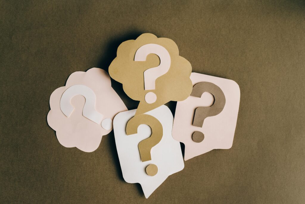 Four paper cutouts with question marks are arranged on a brown surface, each shaped like a speech or thought bubble in beige and light pink tones—perfect for sparking a question of the day for work.