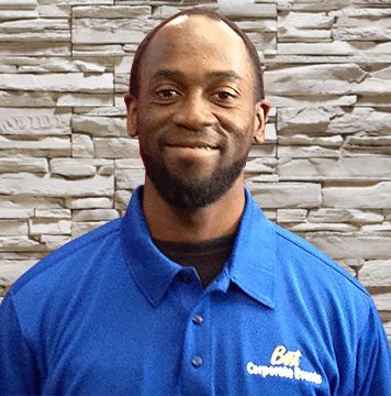 A black man in a blue shirt standing in front of a stone wall.