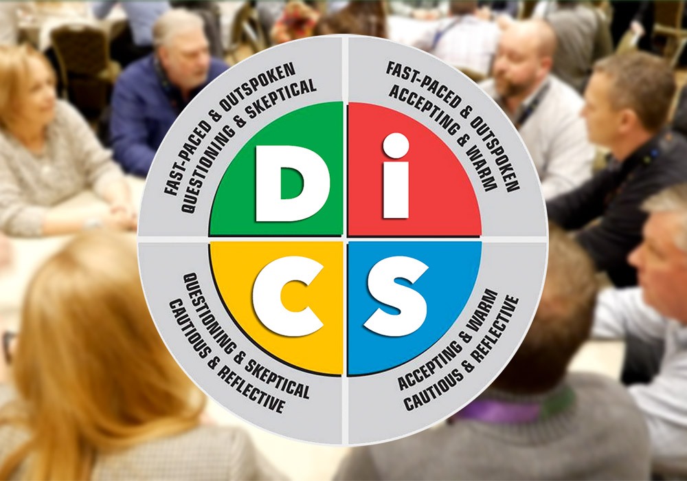 A group of people sits around a table behind a circular DiSC personality chart showing four quadrants with different behavioral traits.