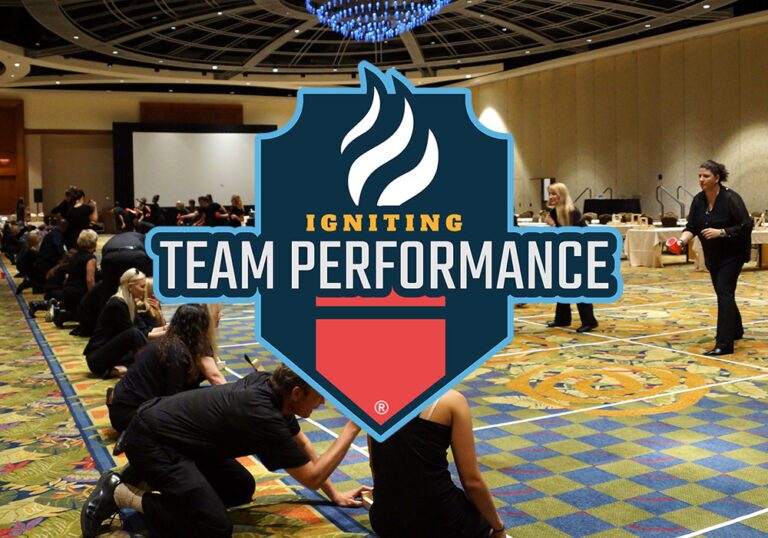 People dressed in black participate in a group activity in a large conference room with a sign reading "Igniting Team Performance" overlaid in the center.