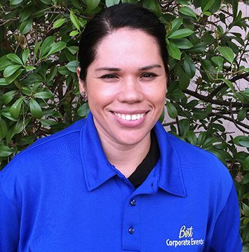 A woman in a blue shirt smiling in front of bushes.