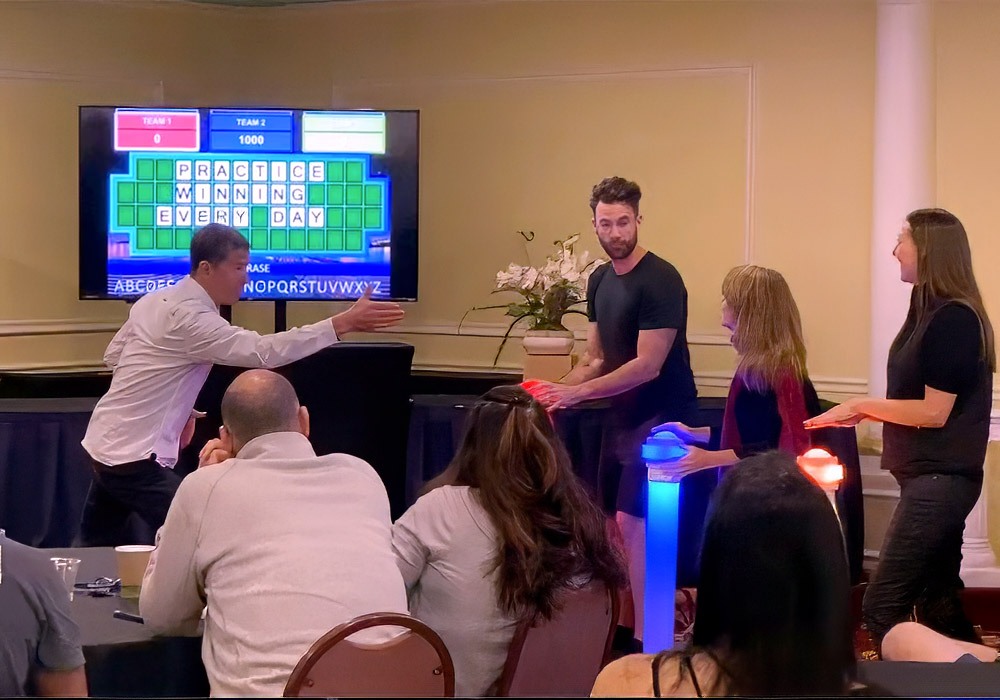 Four people stand at game show-style podiums, engaging in a word puzzle game displayed on a screen behind them. An audience observes the activity from seated positions.