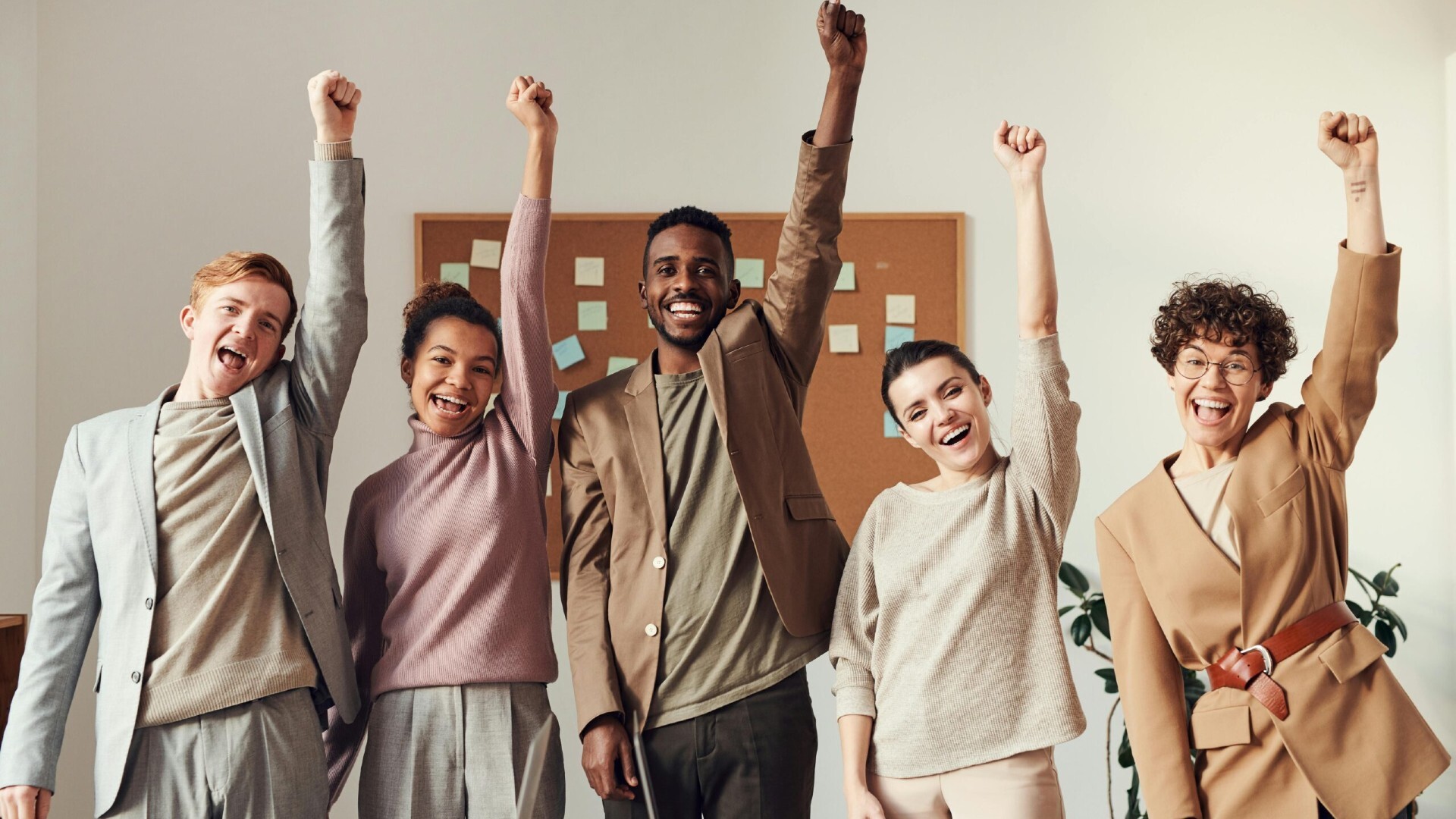 Five people stand side by side indoors, smiling and raising one arm in the air, appearing joyful. A cork board with sticky notes is visible in the background.