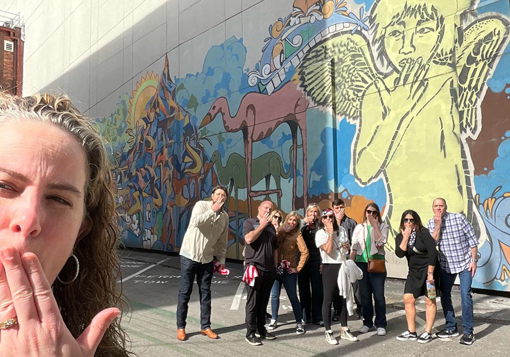 Group of tourists posing for a photo in front of a colorful mural with one person in the foreground blowing a kiss.