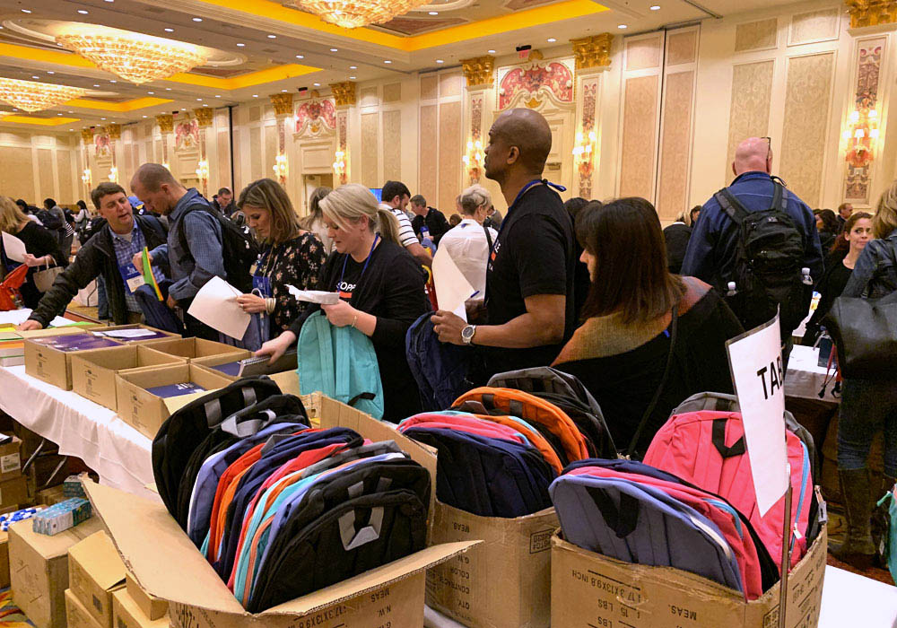 People at a conference stand around tables filled with backpacks and boxes. They appear to be sorting or distributing the items in a large, ornate room.