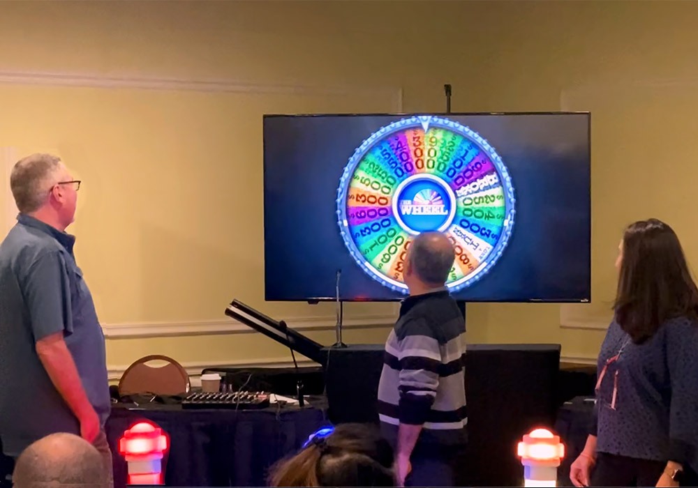 A group of people standing in front of a tv watching a wheel of fortune.