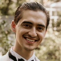 A man with slicked-back hair, a mustache, and a goatee smiles at the camera, wearing a white shirt with a black bow tie. The background is blurred with greenery and a structure visible.
