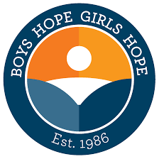 The image is a circular logo for "Boys Hope Girls Hope," established in 1986, featuring an abstract design of a rising sun over a horizon, split into orange and blue sections.