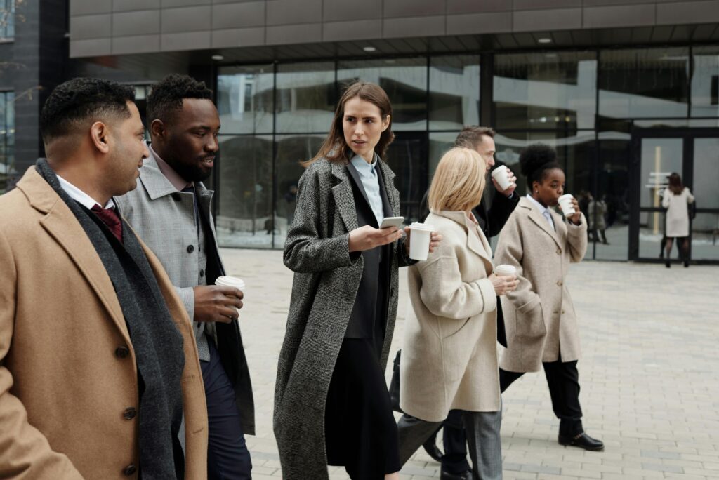 A group of six professionally dressed people walk together outside an office building, each holding a cup of coffee—proof that sometimes coworkers are not your friends, but they do make good company on the way to work.