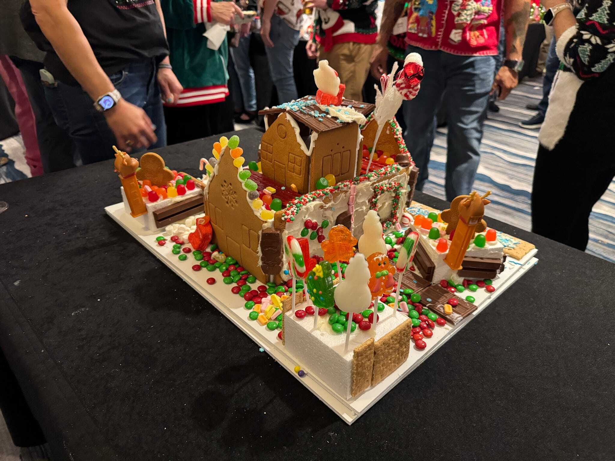 A decorated gingerbread house with candy and cookies sits on a black table, surrounded by people in festive clothing.