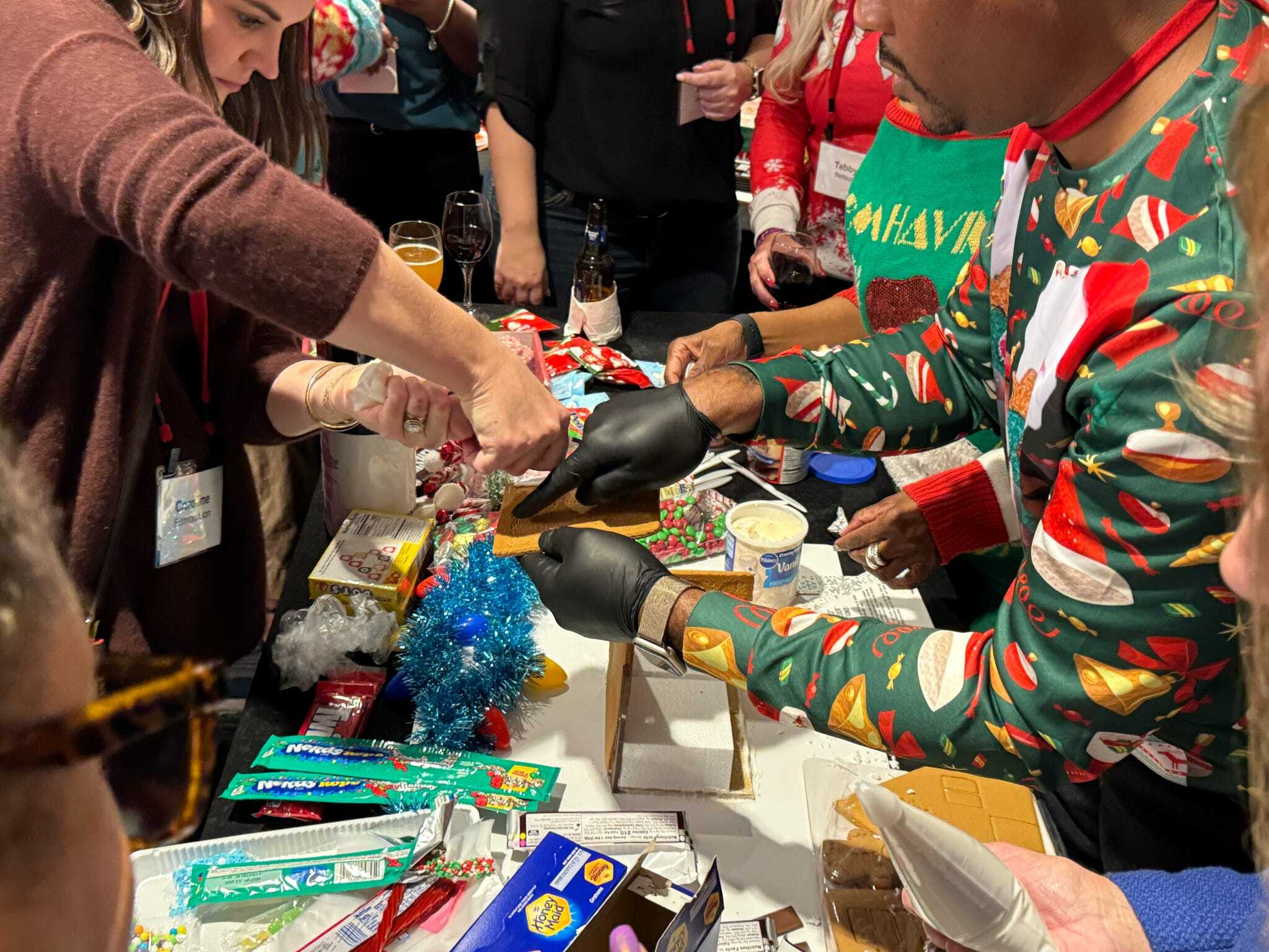 People assemble and decorate gingerbread houses at a festive holiday gathering, using frosting, candy, and various colorful decorations.
