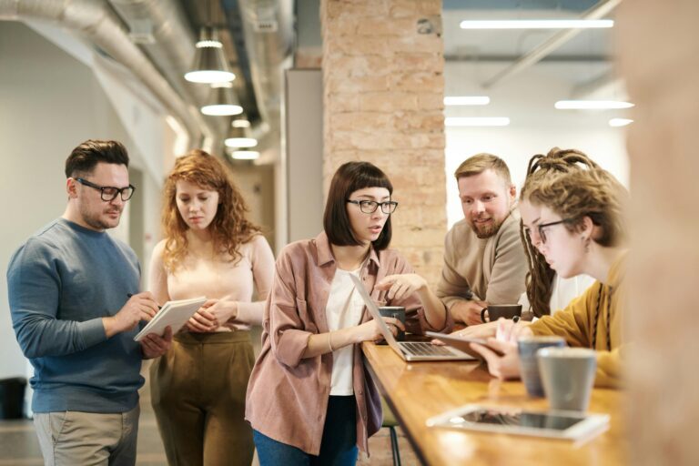 Five Gen Z employees are gathered around a high table in a modern office, discussing something on a laptop and tablets, with coffee cups on the table.