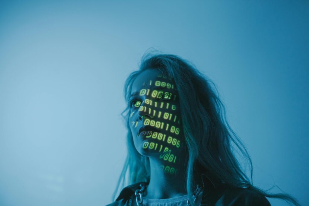 A person with long hair stands in blue lighting as green binary code is projected onto their face, capturing the fear of AI in a digital age.